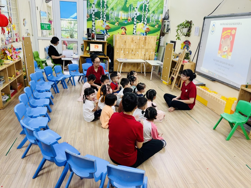 A group of children sitting on the floor in a circle with a person and a projector screenDescription automatically generated